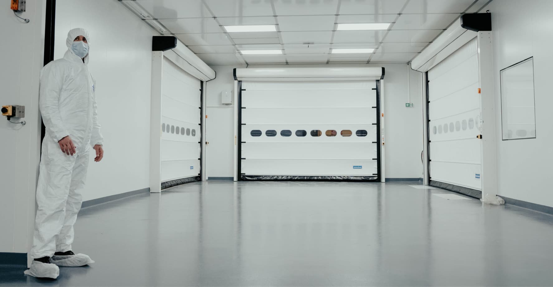 White room with 3 white garage style doors and a person standing wearing hazmat gear